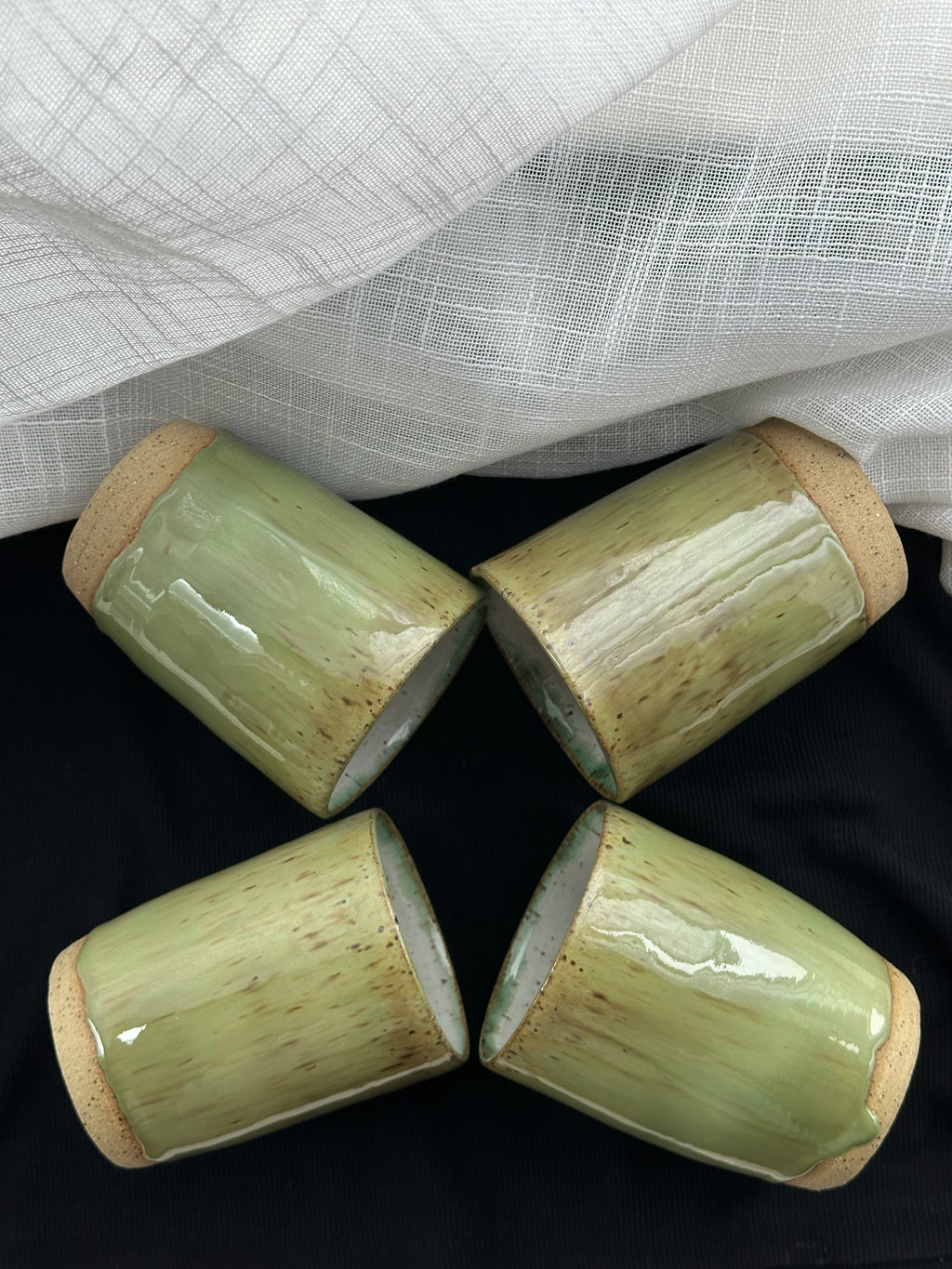 Four cylindrical ceramic cups with a natural finish on a black surface with a white textured fabric background.