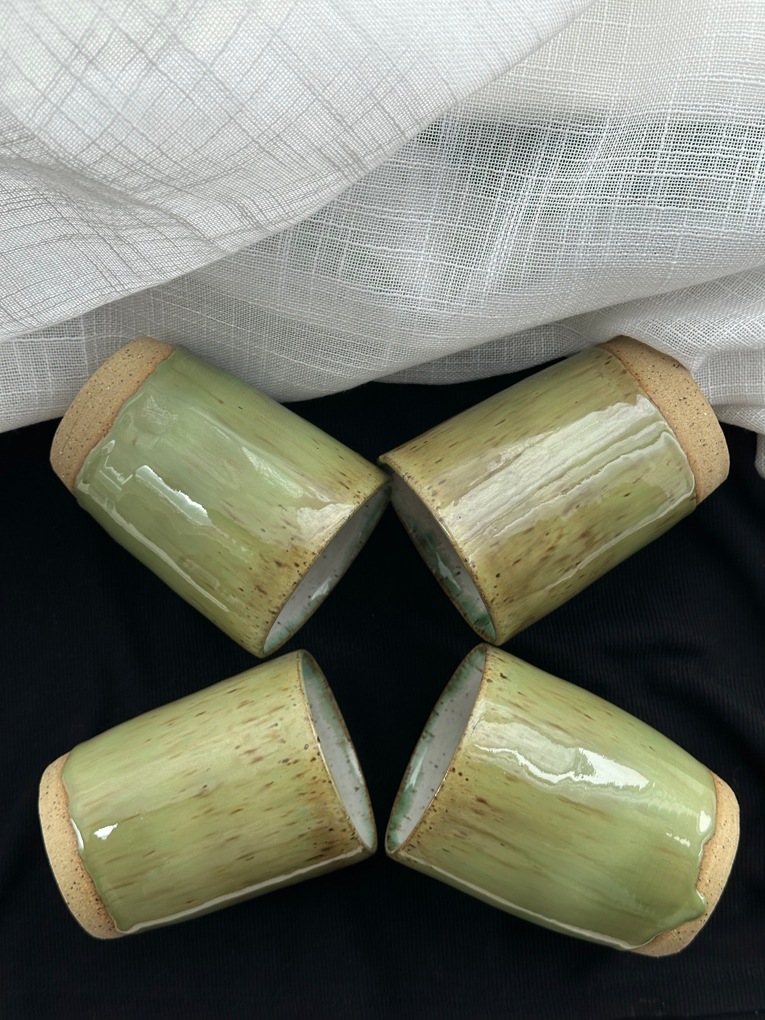 Four cylindrical ceramic cups with a natural finish on a black surface with a white textured fabric background.