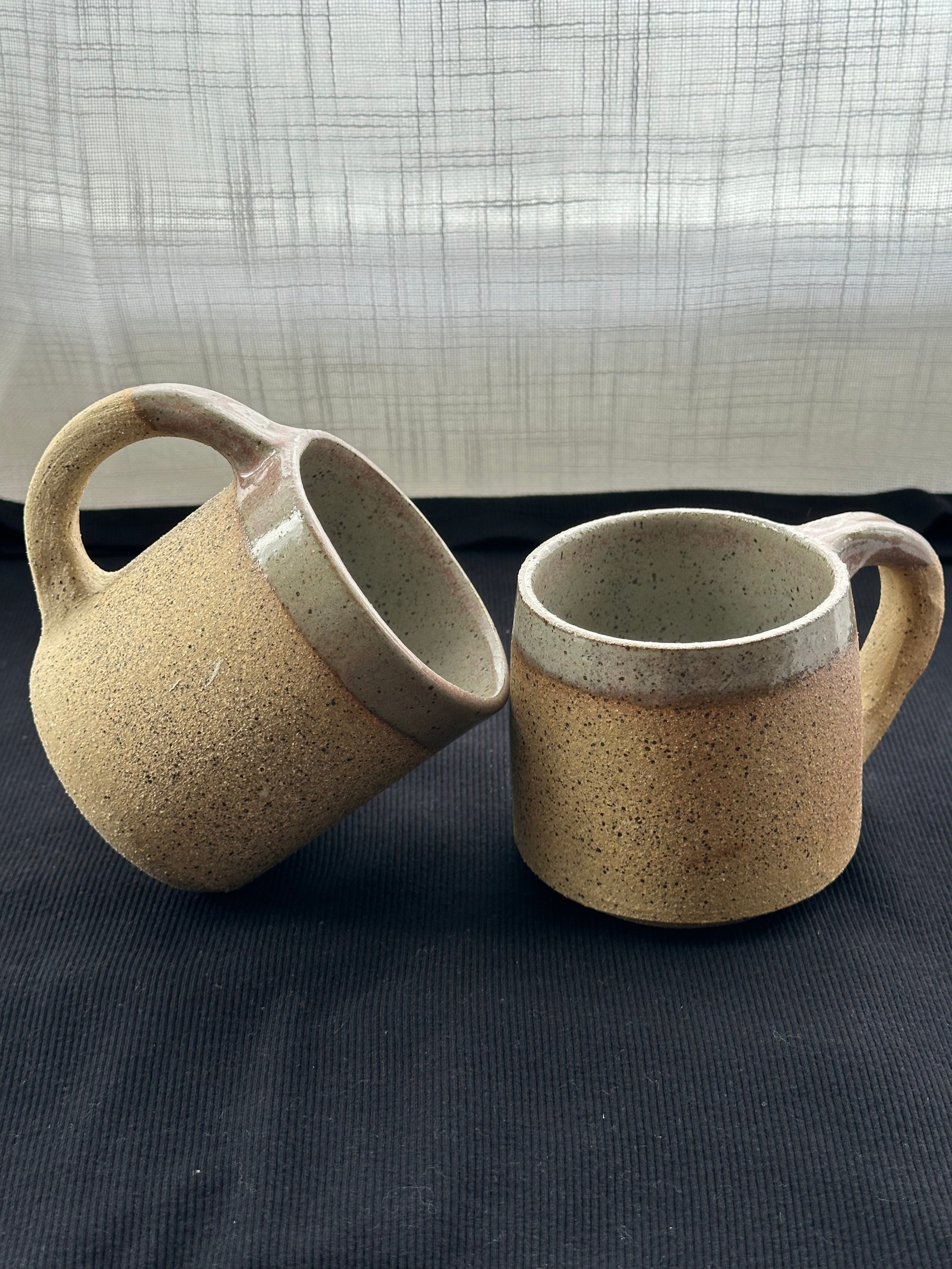 Two ceramic mugs with speckled texture on a dark surface with a light curtain background.